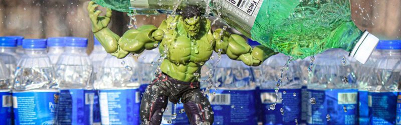 A toy Incredible Hulk smashes a 2 liter of soda while surrounded by many bottles of water.